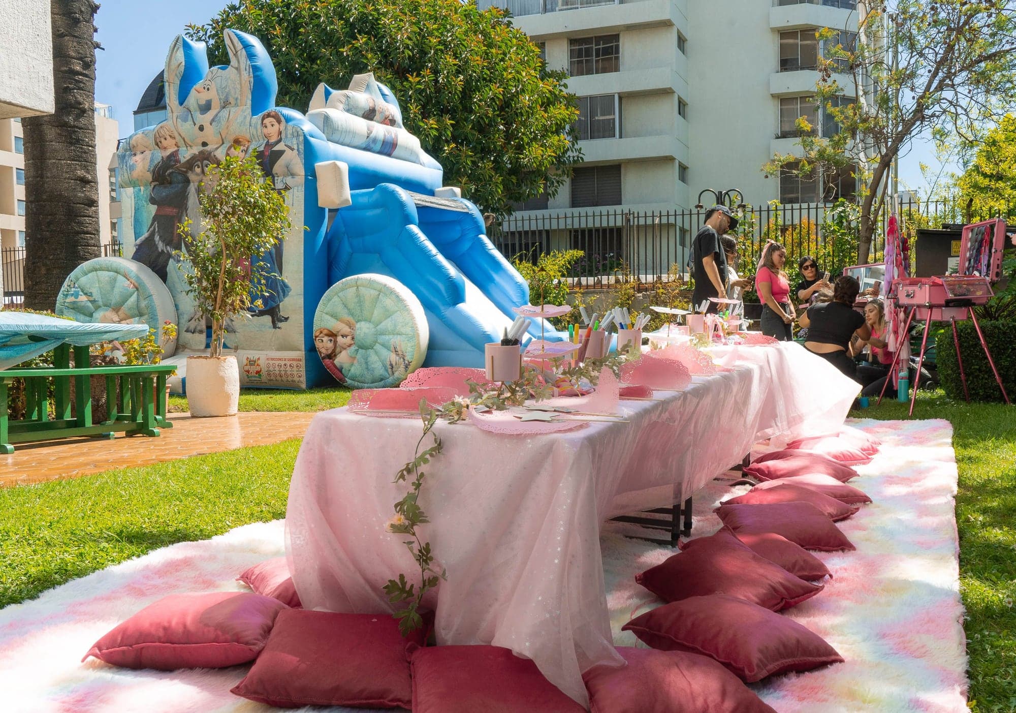 Mesa larga decorada en rosado con cojines sobre el pasto, junto a un inflable
